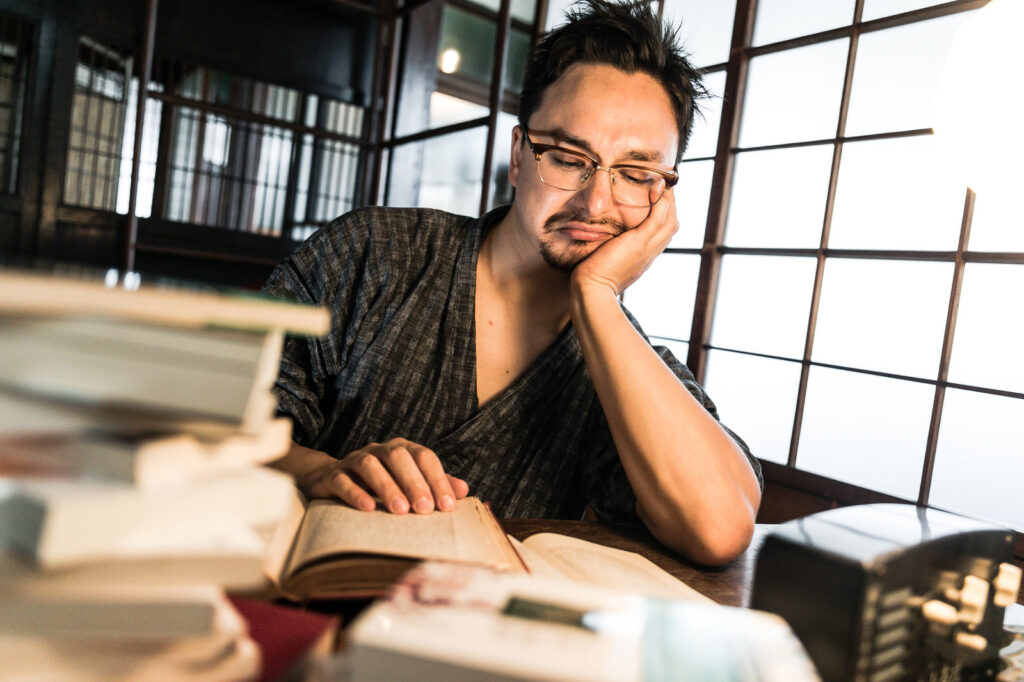 頬杖をつきながら本を読んでいる男性。疲れた表情だが、机には開かれた本と資料が並ぶ。