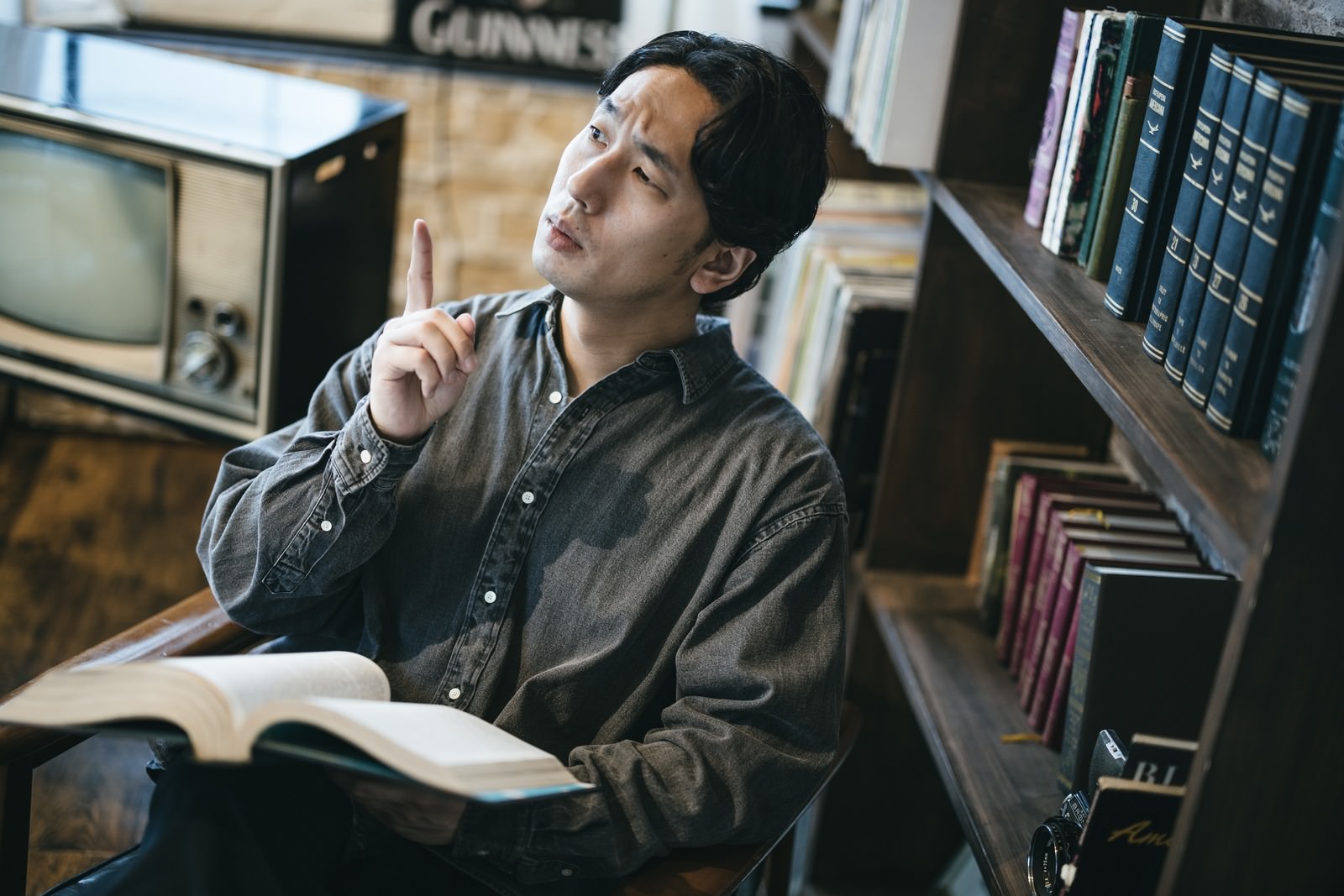 本を開いて指を立てながら考え込む男性。勉強中に思案している様子。